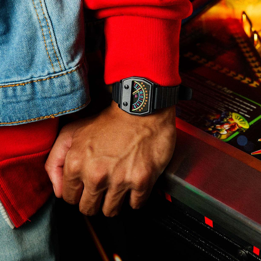 A close-up lifestyle shot of a person wearing a rectangular black watch with a red and blue display, resting their hand near a pinball machine.
