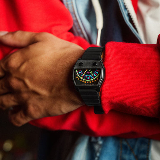 A close-up of a person's wrist wearing a black Sector watch with a colorful roulette-style face, styled with a red hoodie and denim jacket against a dark, blurred bokeh background.