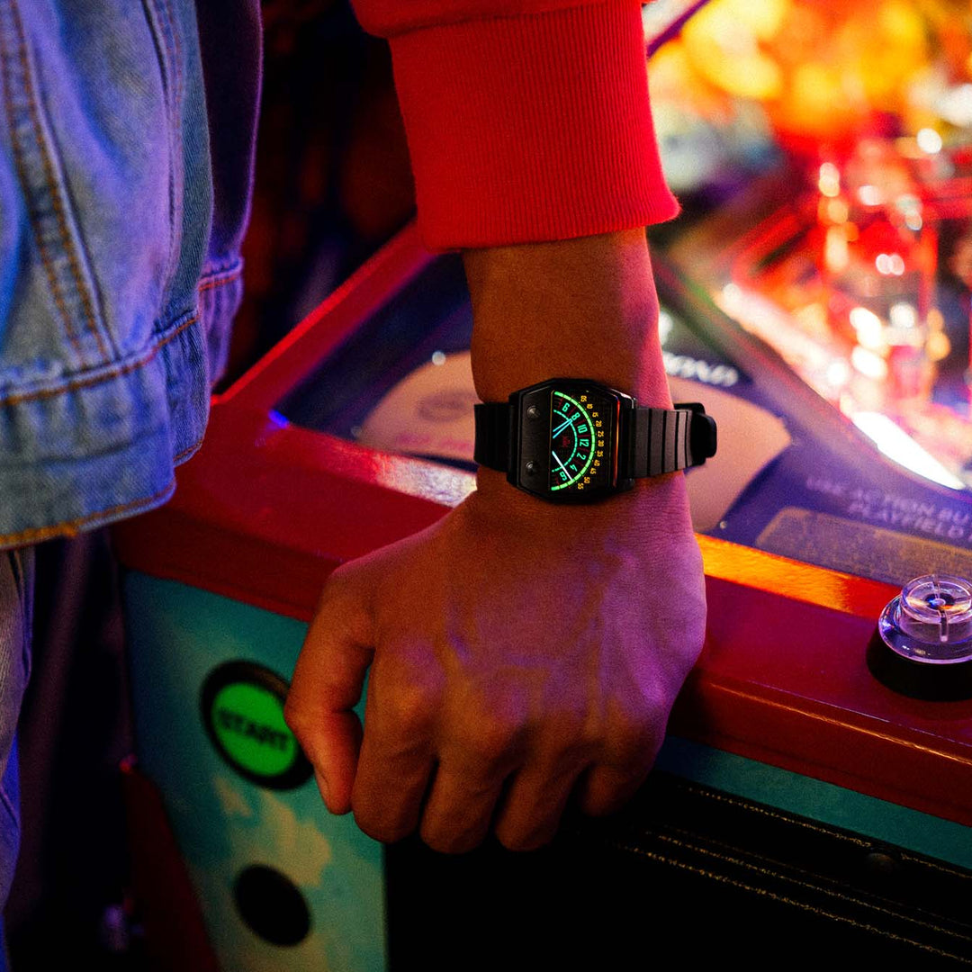 A close-up lifestyle shot of a person's hand resting on an arcade machine, wearing a black digital wristwatch with a glowing green display.