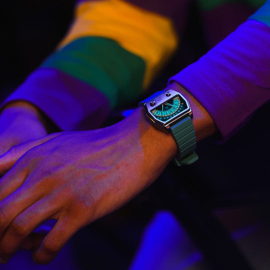 A close-up of a person's hands and arms wearing a green smartwatch with a green band, styled with a colorful striped long-sleeve shirt under dramatic purple and blue lighting.