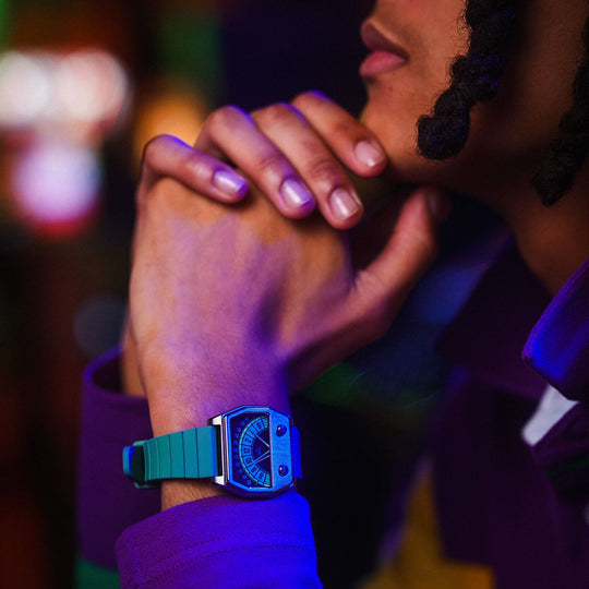 A close-up lifestyle shot of a person wearing a teal smartwatch with a square silver case and a dark strap, set against a dark, colorful background with purple and green lighting.