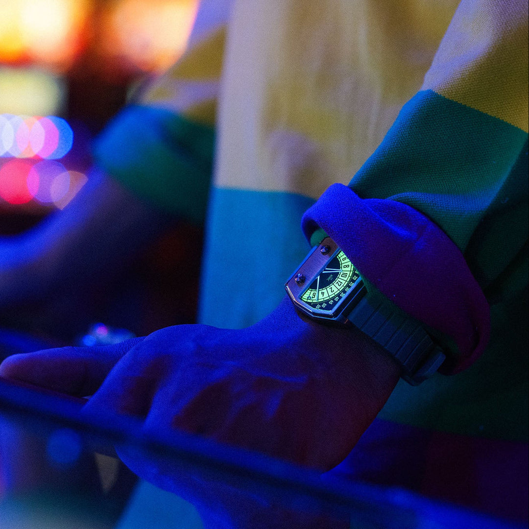 A close-up lifestyle shot of a person's hand and wrist wearing a smartwatch with a glowing green face and a purple sleeve, set against a blurred background of colorful arcade lights.
