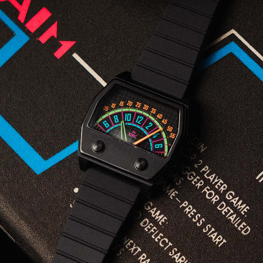 A black wristwatch with a retro, colorful digital game-style face and black strap, styled diagonally on top of a vintage arcade machine control panel.