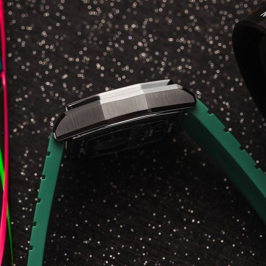 A close-up shot of a smartwatch with a brushed metal case and a green textured strap, resting on a black speckled surface next to a colorful object and a camera lens.