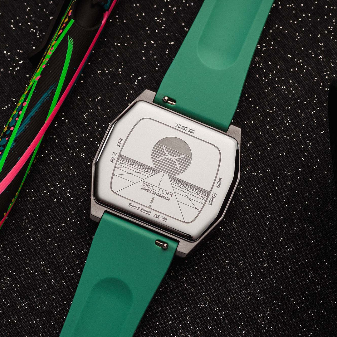 A close-up of a wristwatch with a bright green silicone strap and a silver-toned case, shown from the back on a dark textured surface. A colorful strap or band lies to the left, and part of a black lens is visible on the right.