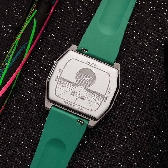 A close-up of a wristwatch with a bright green silicone strap and a silver-toned case, shown from the back on a dark textured surface. A colorful strap or band lies to the left, and part of a black lens is visible on the right.