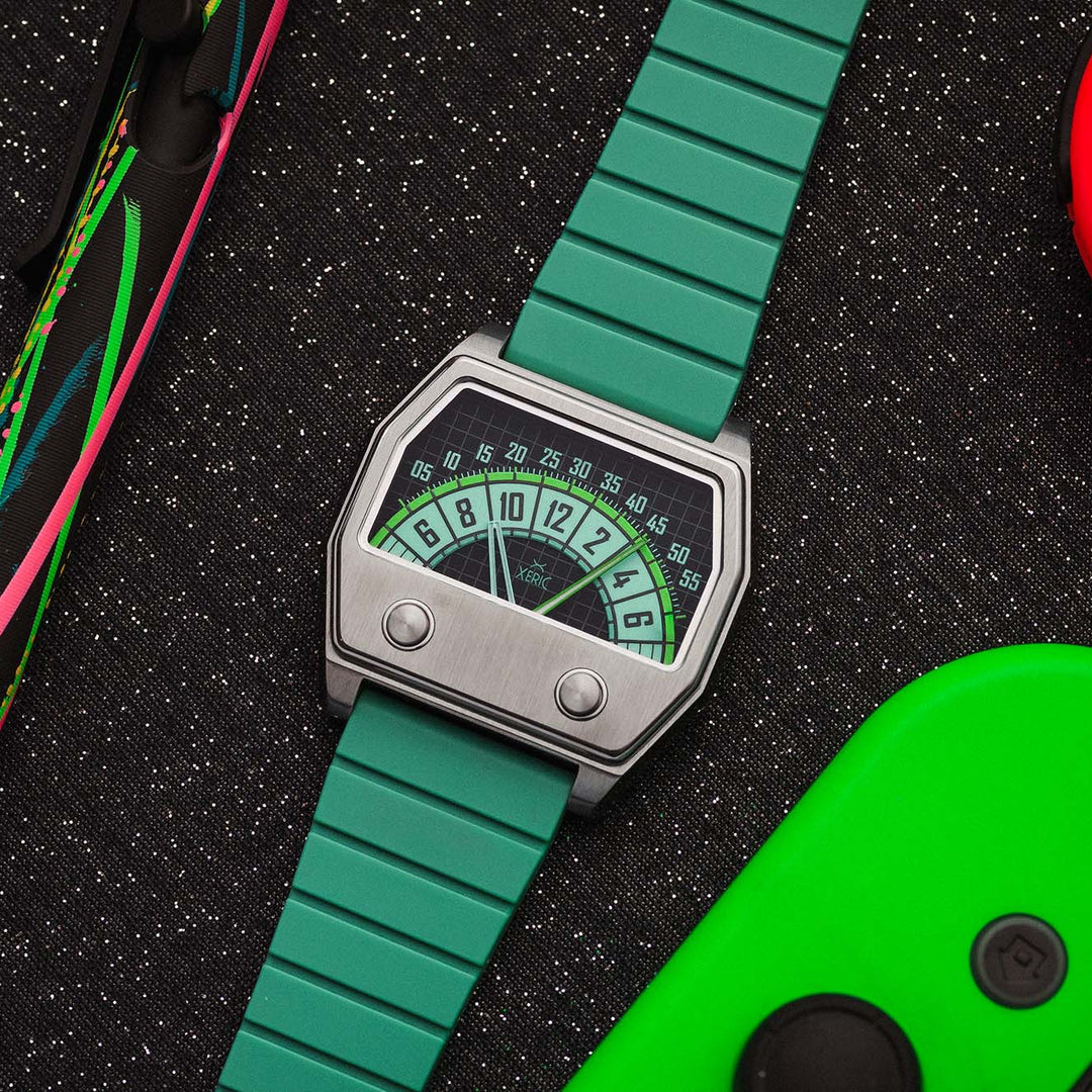 A wristwatch with a bright green segmented strap and a silver-toned rectangular case featuring a retro dial face, styled on a black textured surface with colorful game controller parts nearby.