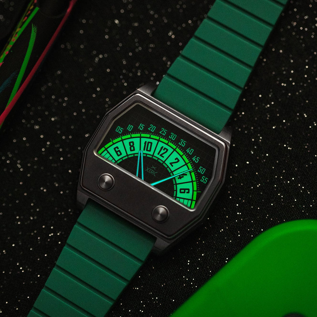 A retro-style wristwatch with a green segmented strap and a dark gray rectangular case, featuring a glowing green analog dial face. The watch is photographed on a black textured surface with colorful abstract props in the corners.