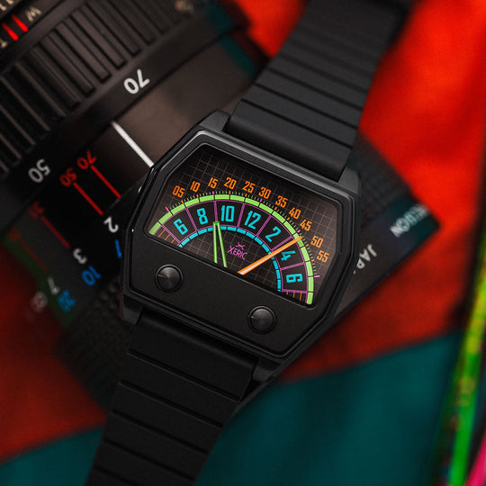 A black analog watch with a colorful retro dial, styled diagonally across a camera lens and a bright multicolor fabric background.