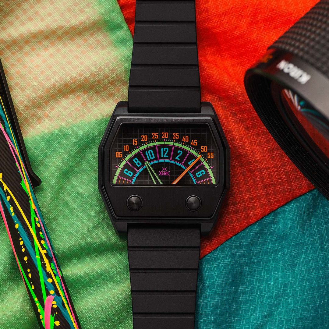 A black digital wristwatch with a colorful retro gauge face, styled on a vibrant multicolor fabric background alongside a pen and a camera lens.