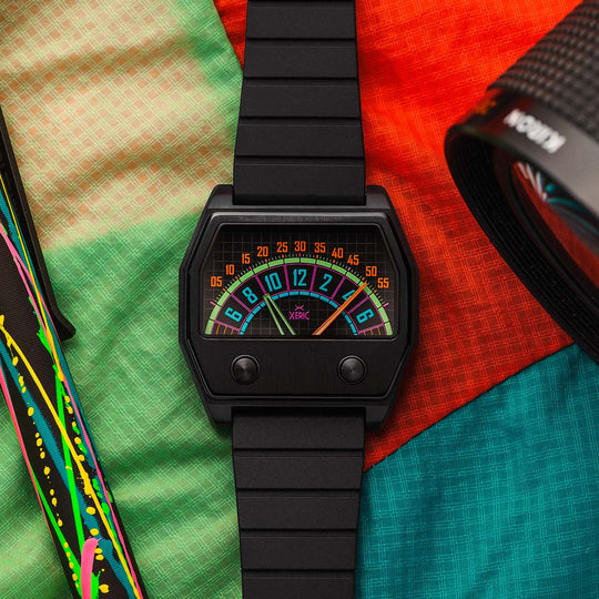 A black digital wristwatch with a colorful retro gauge face, styled on a vibrant multicolor fabric background alongside a pen and a camera lens.