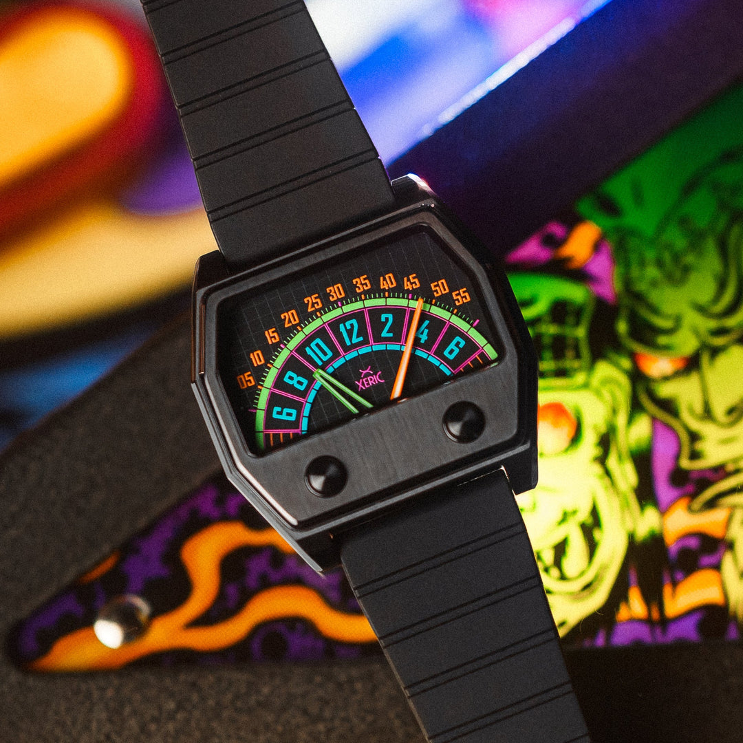A black analog watch with a segmented strap and a colorful, retro-style dial featuring neon green, orange, and blue markings, shown close-up against a vibrant, blurred background.