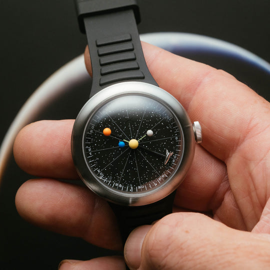 A close-up of a wristwatch with a black strap and a detailed solar system face, held in a person's hand against a dark background.