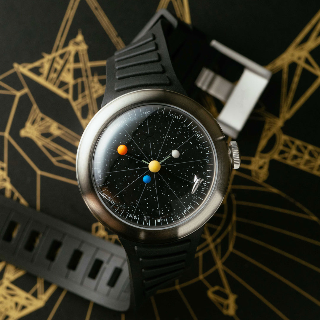 A wristwatch with a black rubber strap, a brushed silver-tone case, and a detailed solar-system-themed dial featuring a black starry background with colorful sun-like planets. The watch is photographed on a dark surface with gold geometric line art.