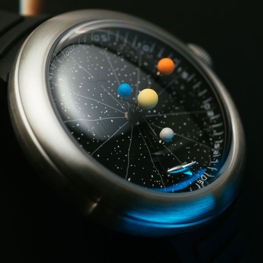 Close-up of a wristwatch with a brushed silver metal case and a black dial featuring a solar system design with colorful planet markers.