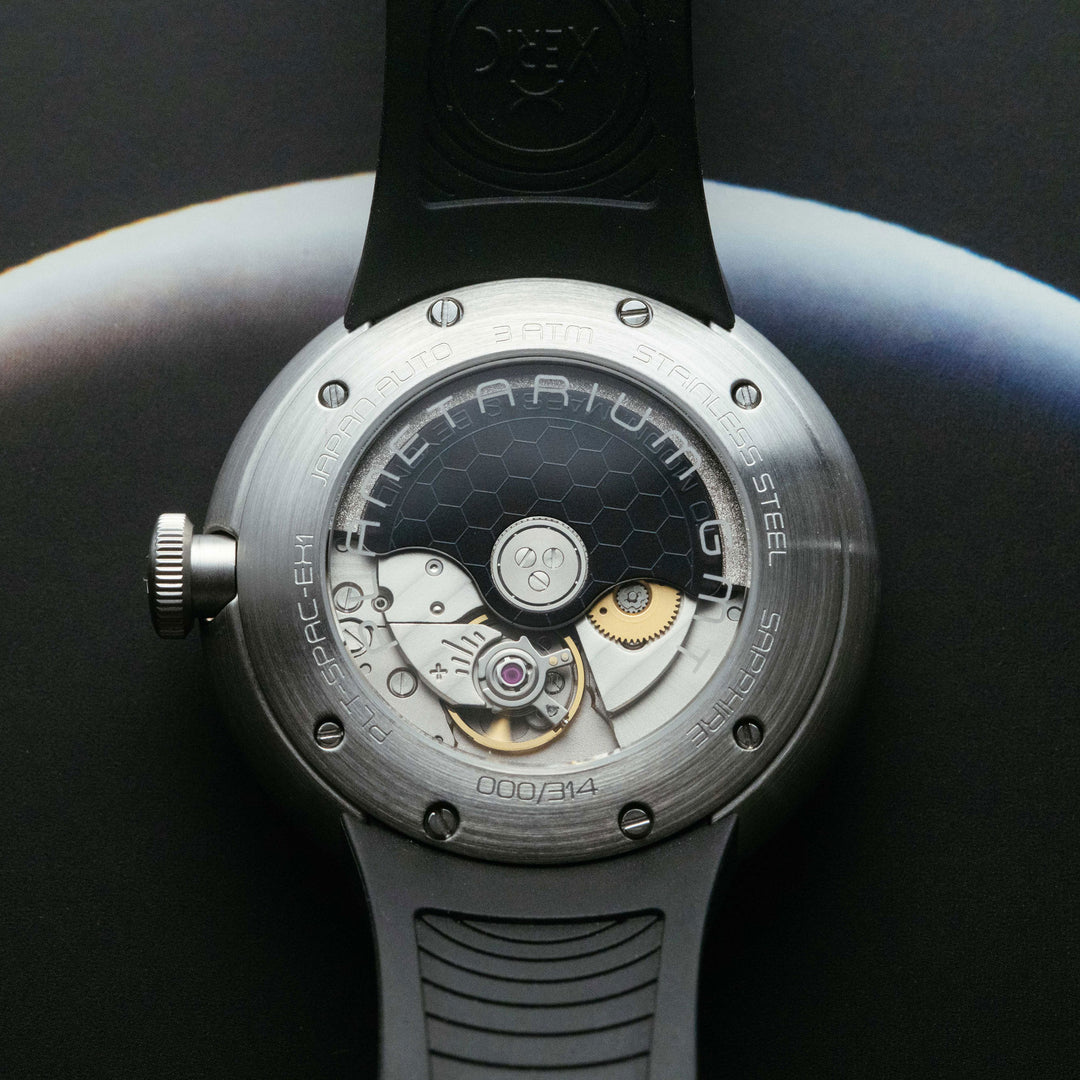 Close-up of a wristwatch with a transparent round case showing the internal mechanical movement, a black dial, and a black strap on a dark studio background.